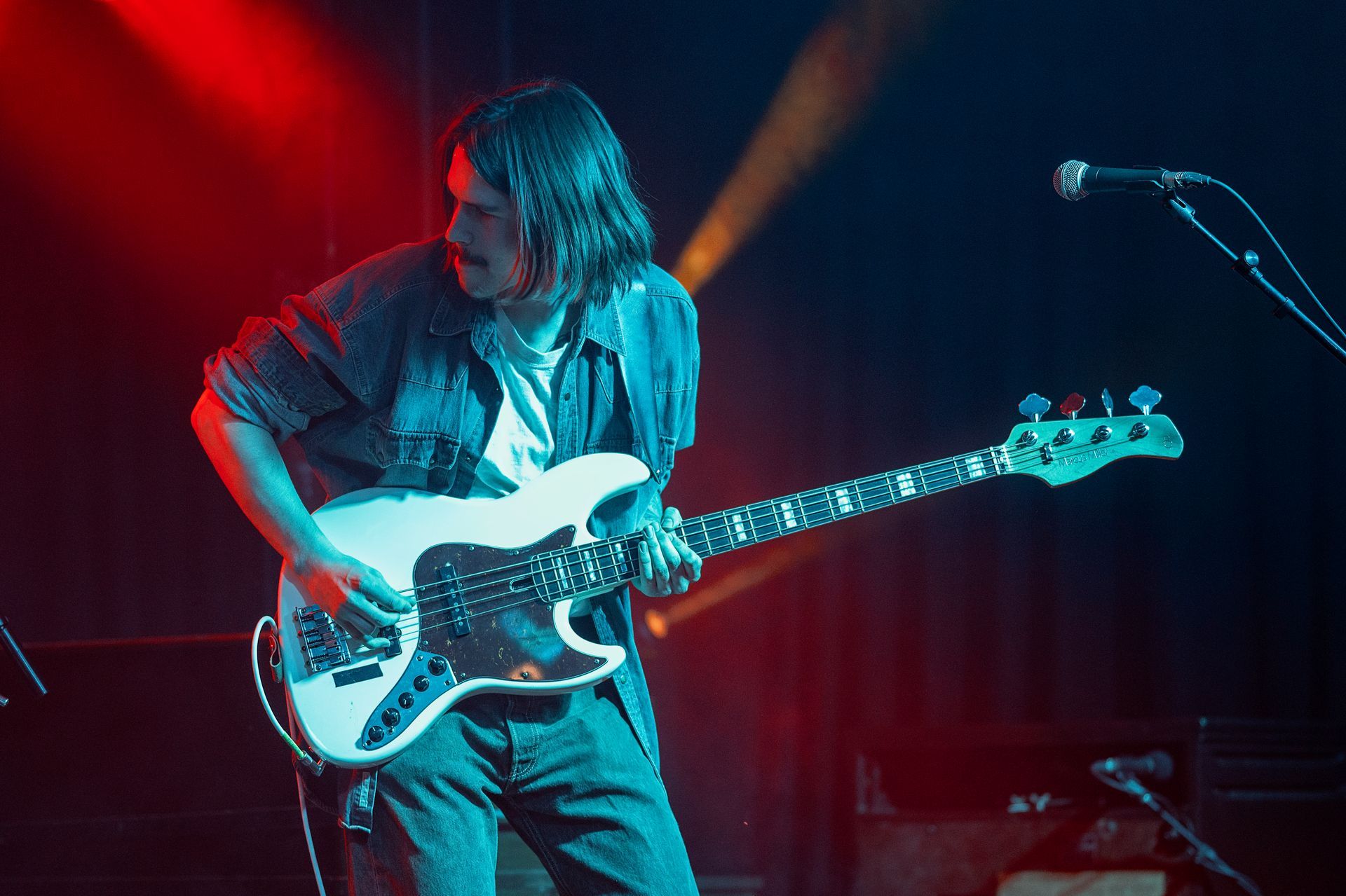Man playing white bass guitar on stage under red and blue lights.