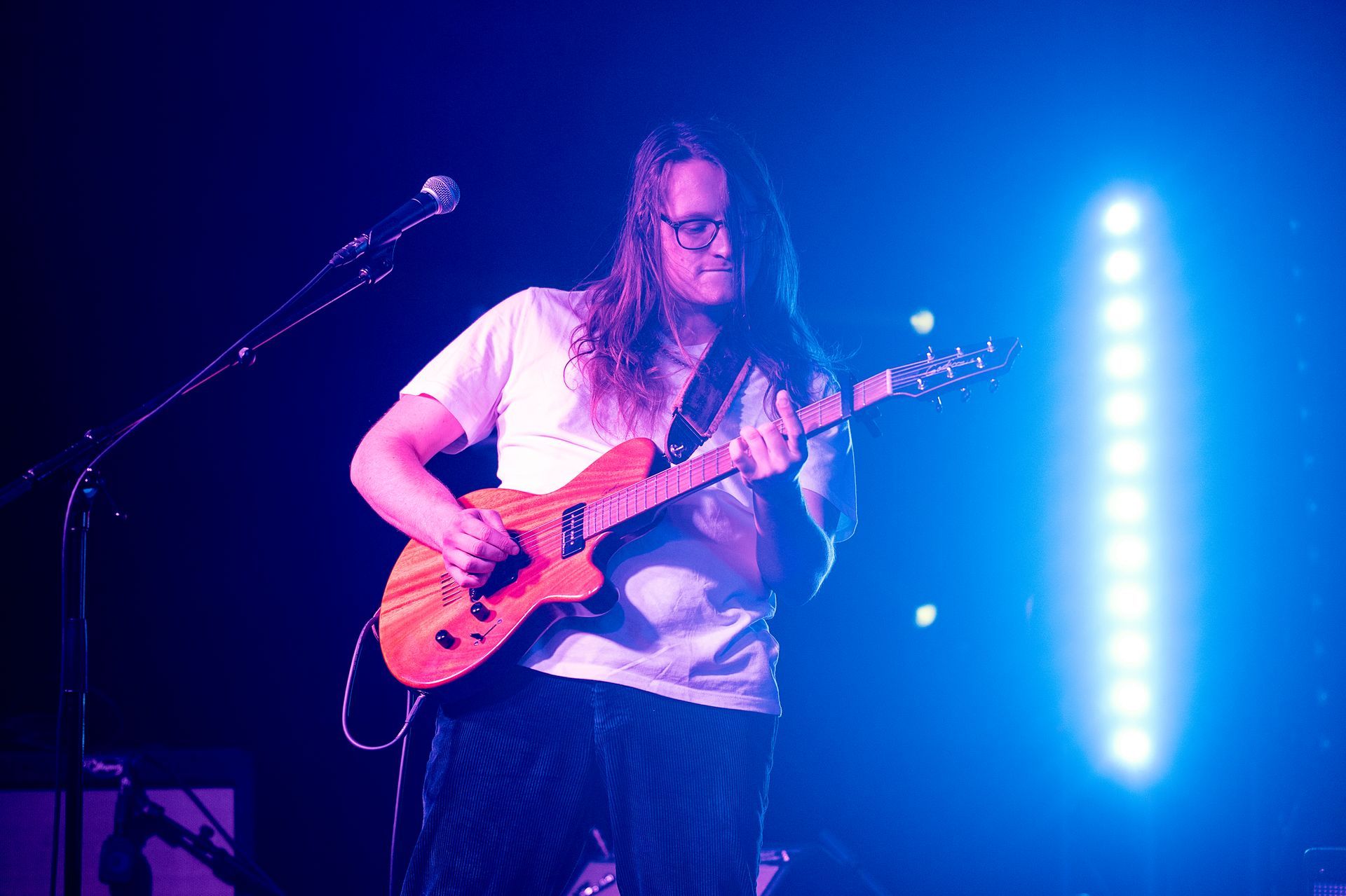 A person with long hair plays an electric guitar under blue stage lights.