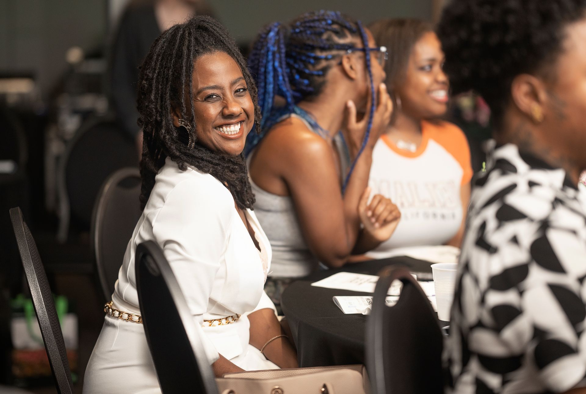 Woman in white jacket smiles at the camera, seated at a table with other attendees at an event.