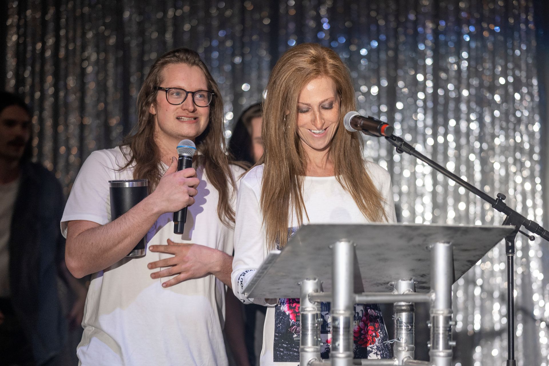 Two people at a podium with microphones, silver backdrop. Woman in white reading, man in white holding cup.