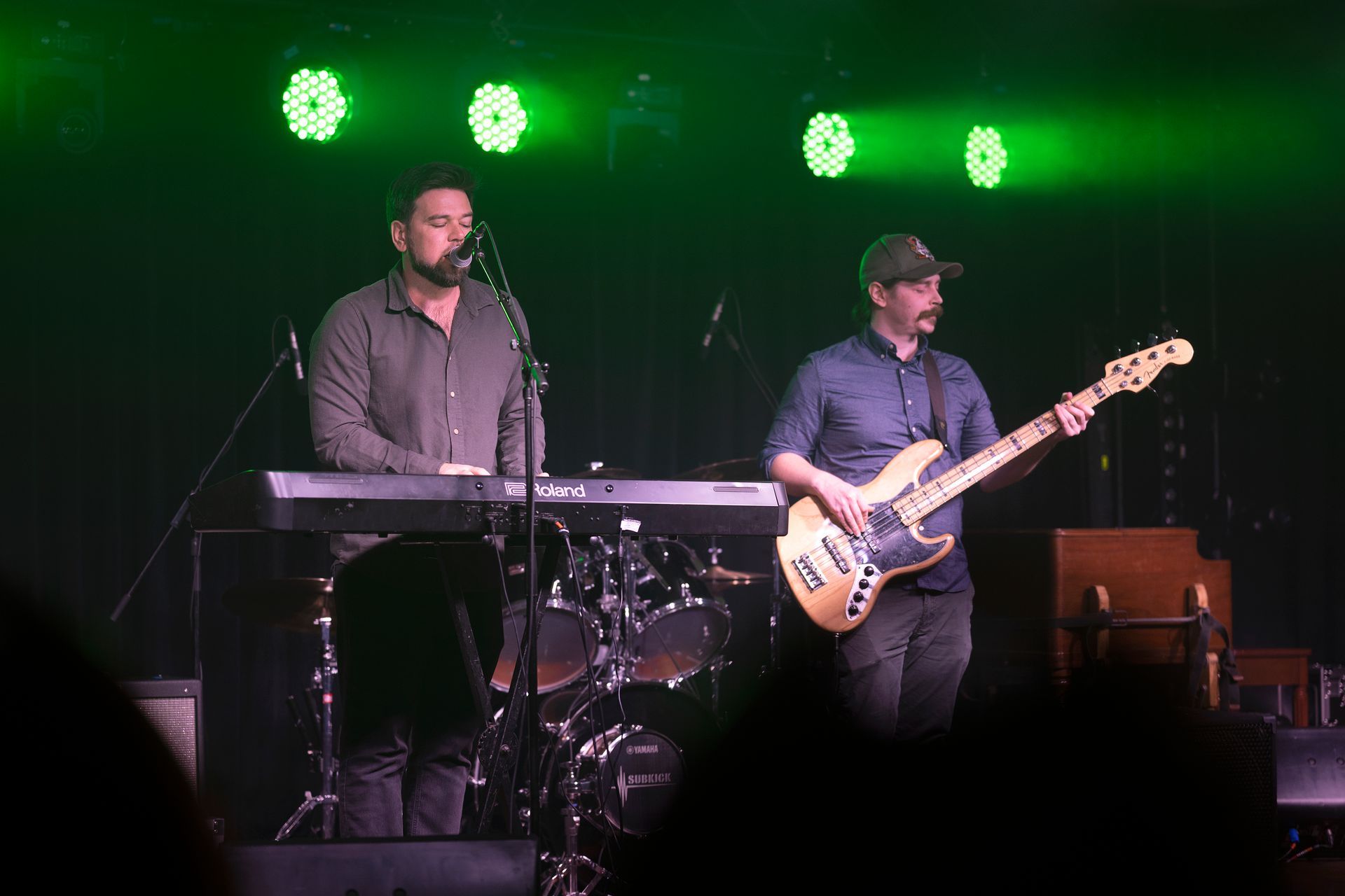 Two men on stage performing: one plays keyboard, the other bass guitar, under green stage lights.