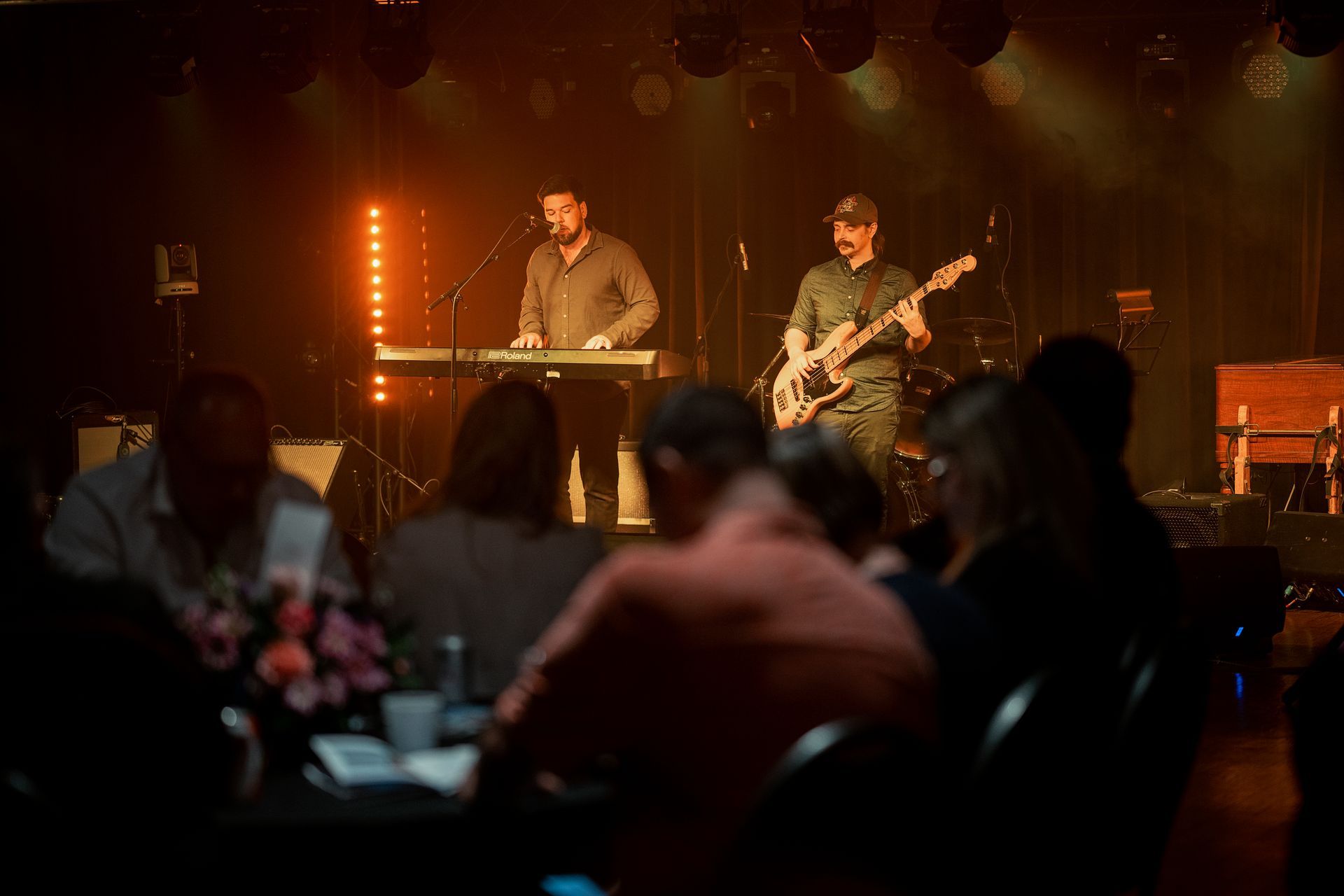 Band playing on stage with audience in foreground. Warm stage lighting.
