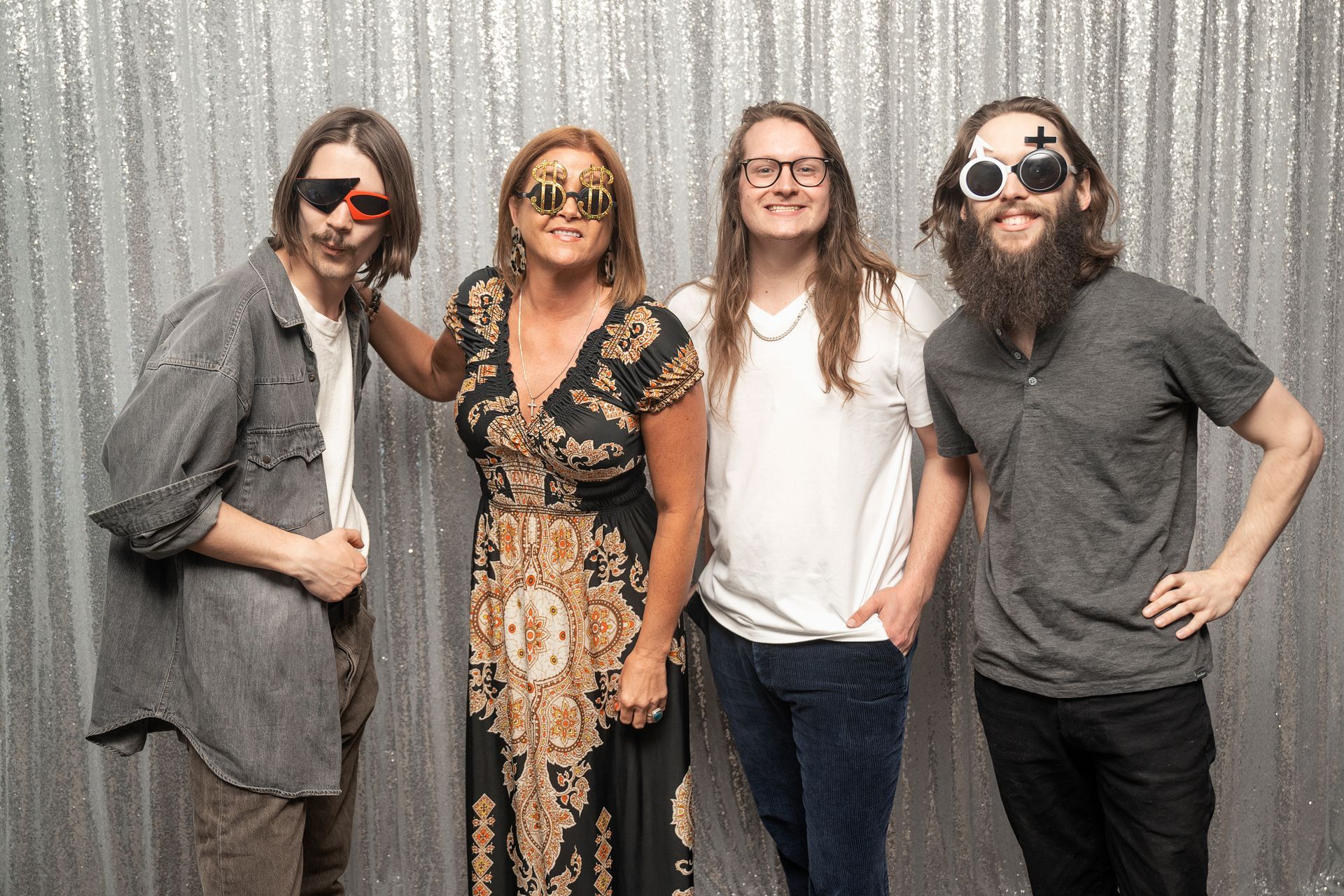 Four people posing in front of a silver backdrop; all wearing novelty glasses; woman in patterned dress; man with beard.