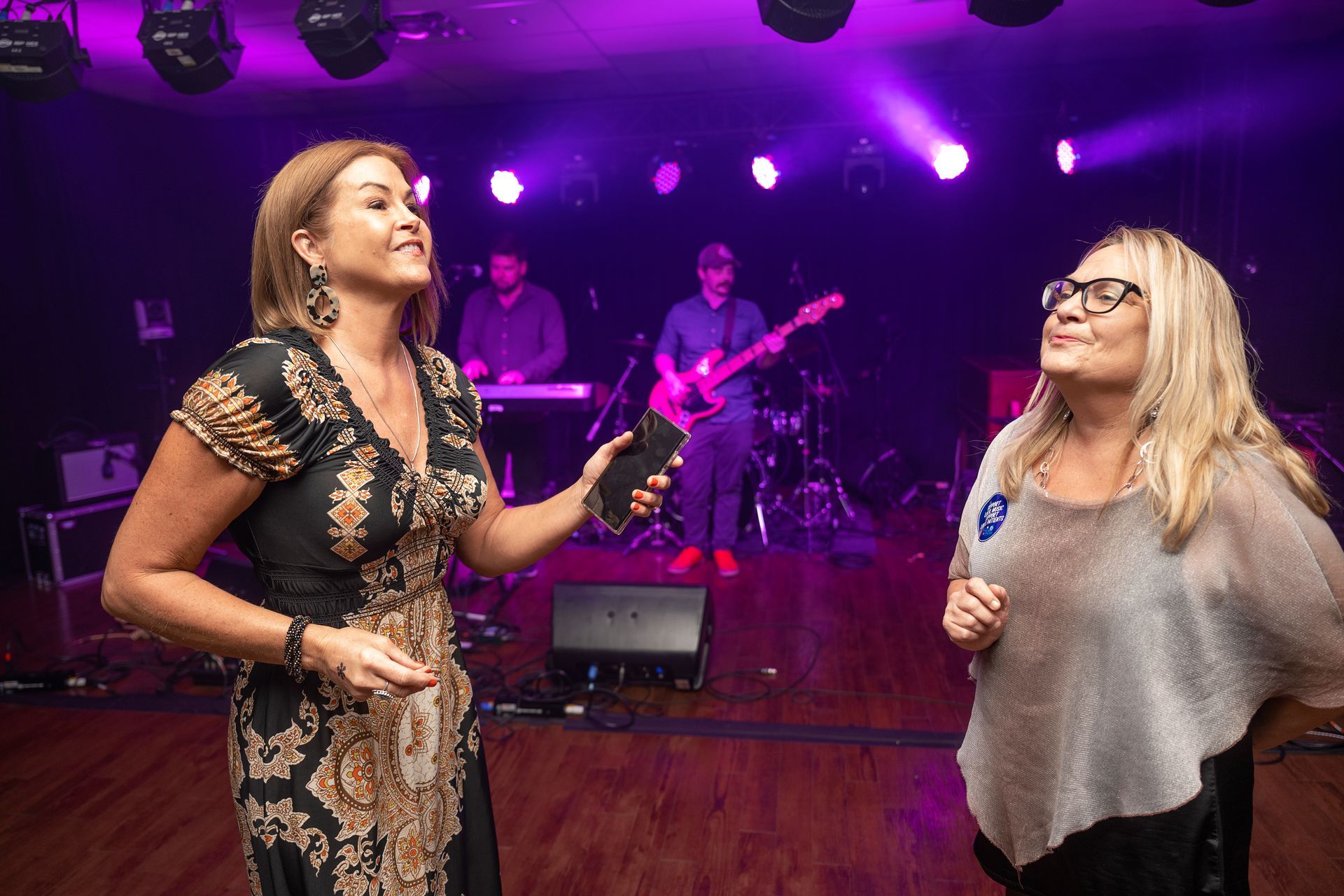 Two women on stage with a band, one holding a phone. Purple lighting.