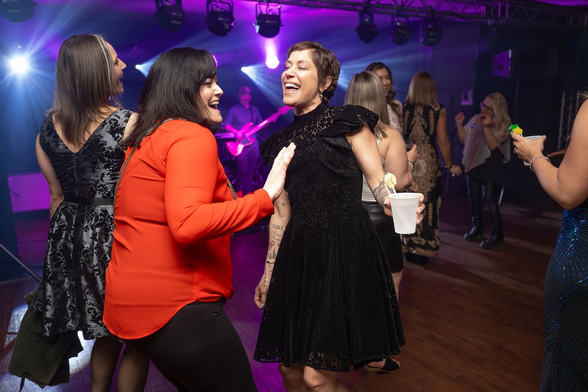 People dancing at a party in a dimly lit room with a band in the background. Two women laugh, one with arms out.