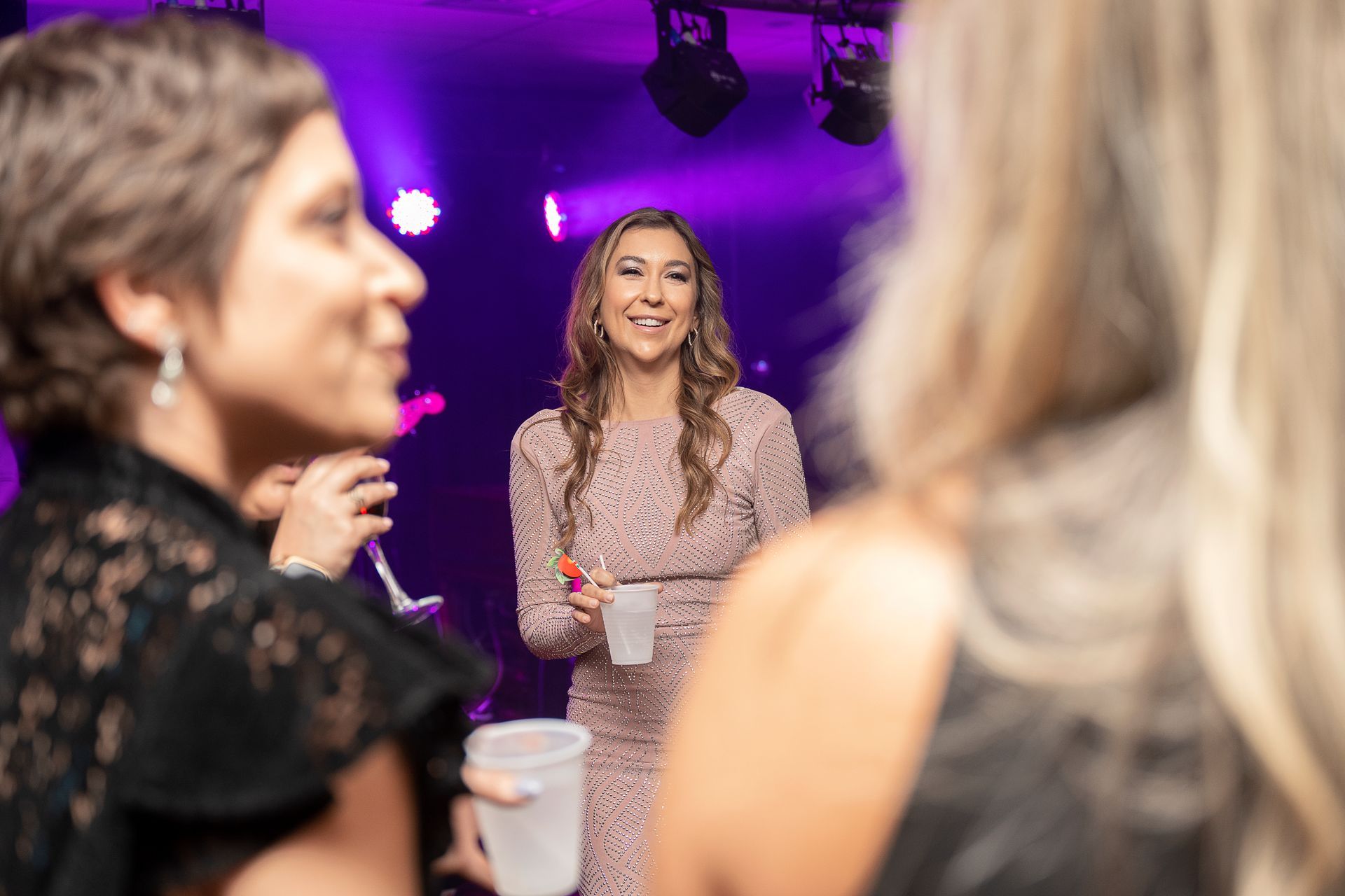 Woman in pink sequined dress smiles, holding a drink, at an event with friends.