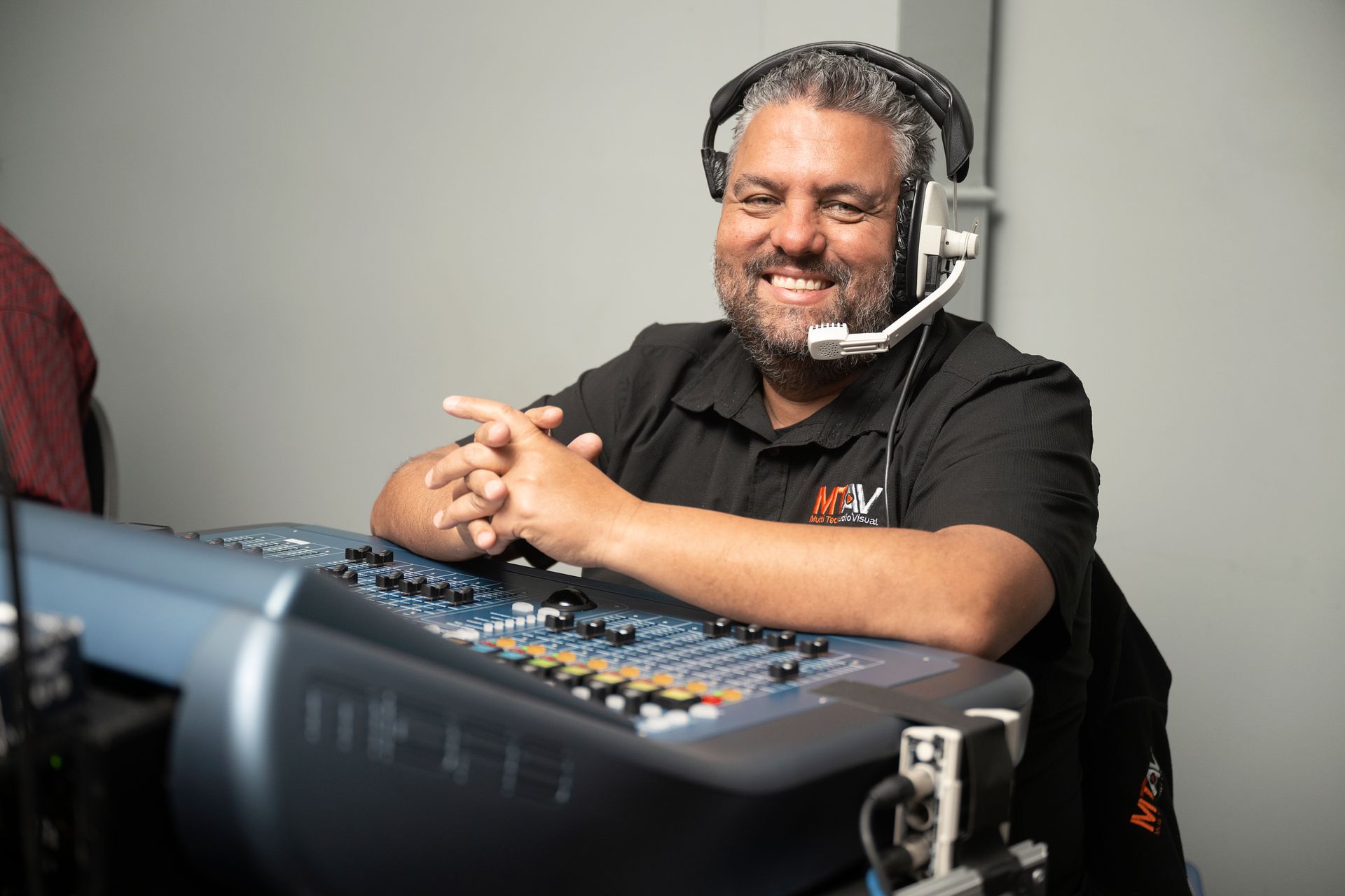 Man wearing a headset smiles while leaning on a sound mixing console. He's in a studio, wearing a black shirt.
