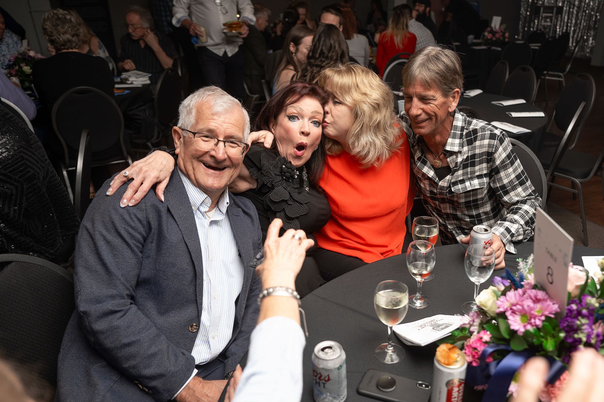 Group of people at a table, smiling, laughing, embracing. A woman kisses another woman's cheek. Setting is a social gathering.