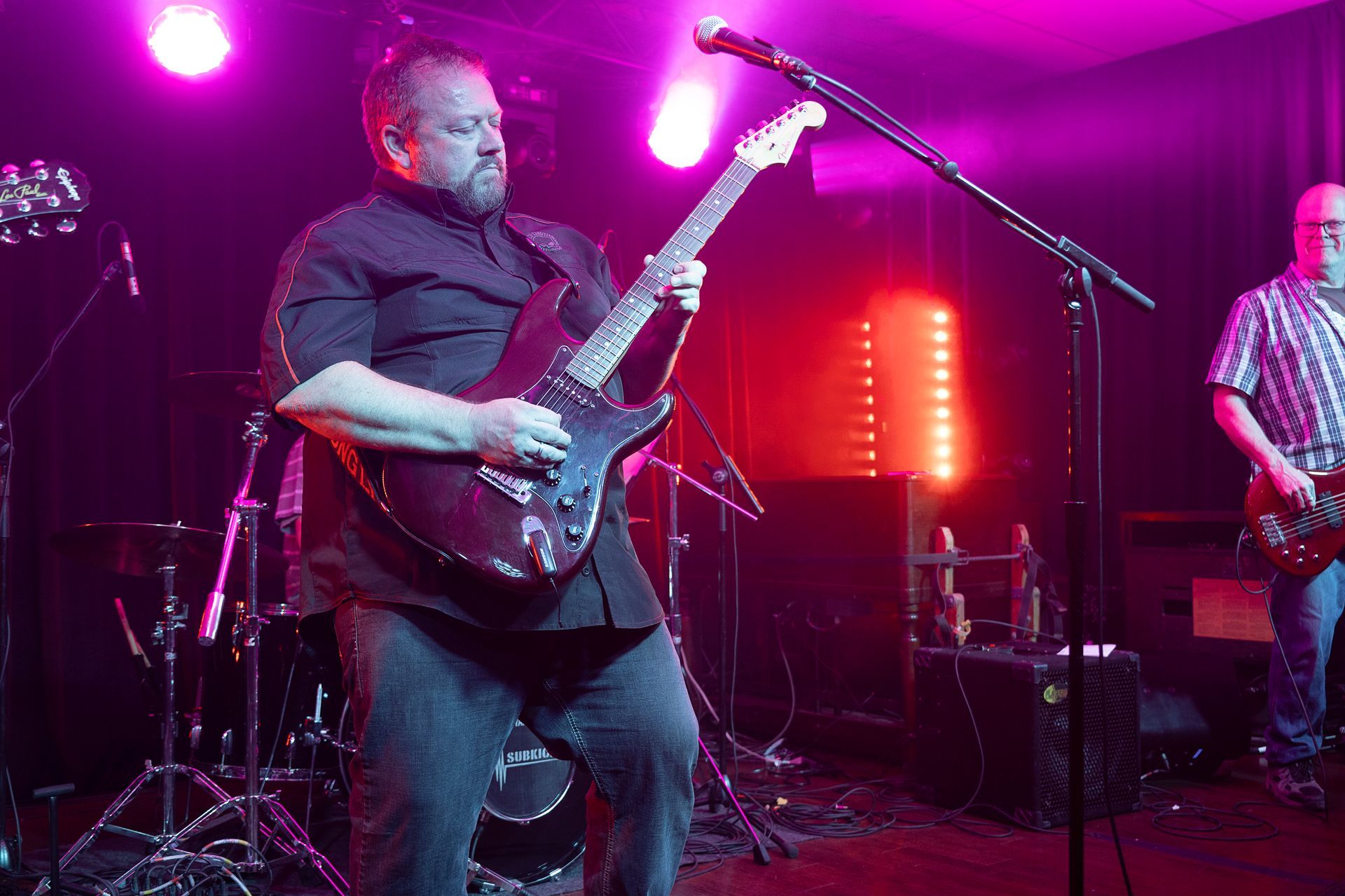 Guitarist on stage, playing, lit with pink lights, another musician in background.