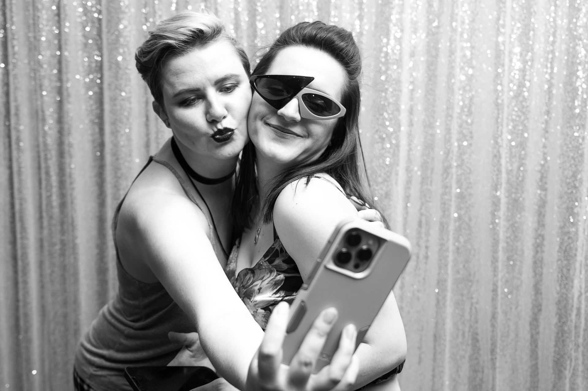 Two people taking a selfie in front of a sparkly backdrop. One pouts, the other wears sunglasses.