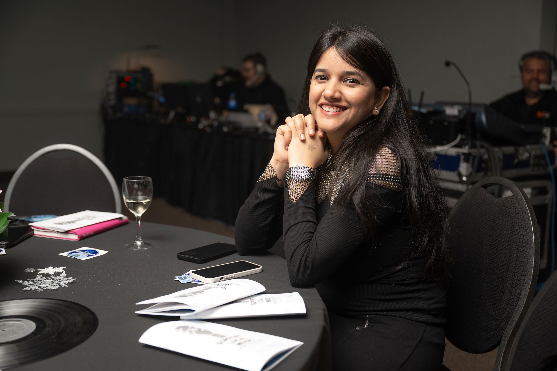 Woman smiling at a table with champagne, phones, and papers. Sound equipment in the background.