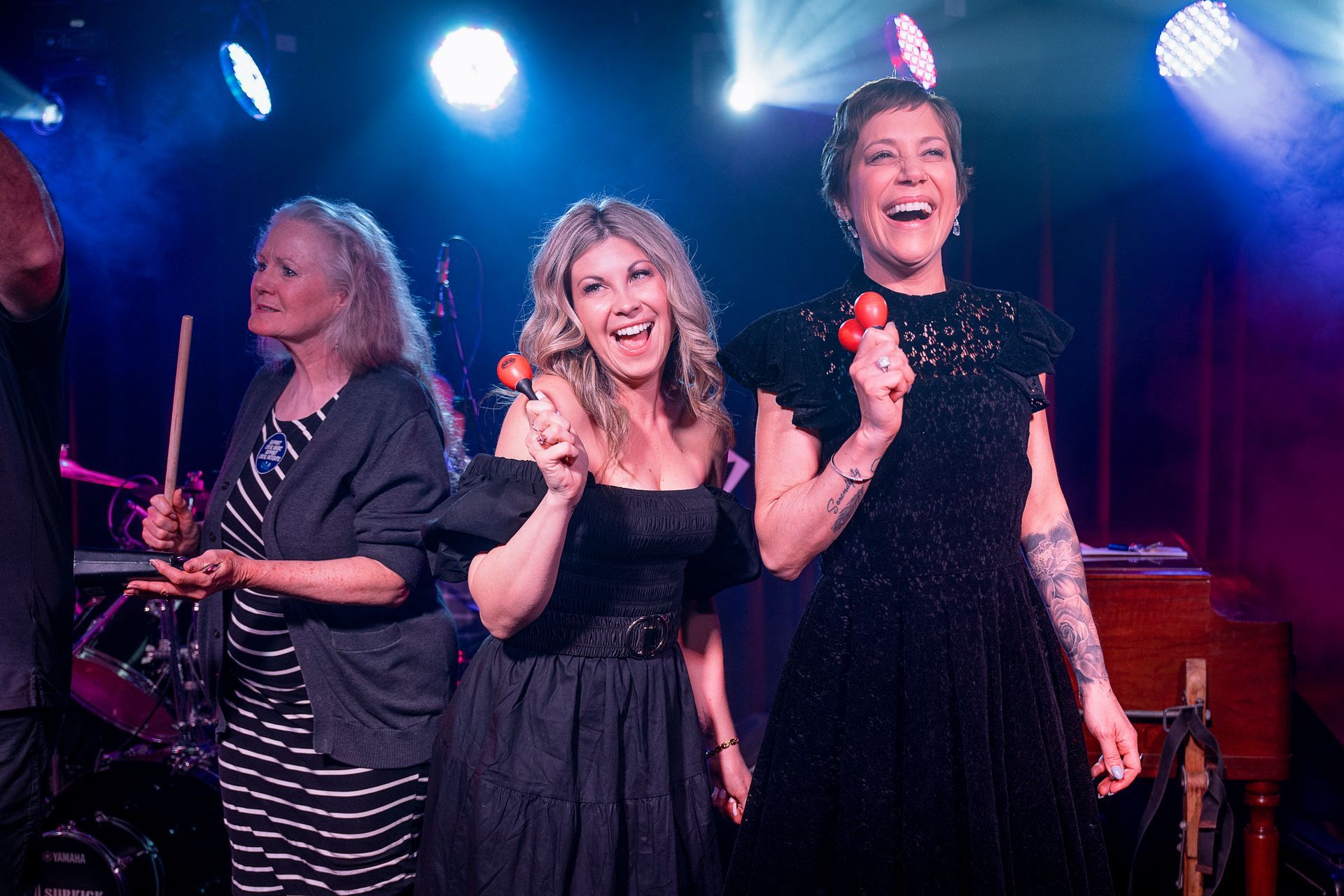 Three women on stage, smiling and holding maracas. Stage is lit with blue and pink lights.
