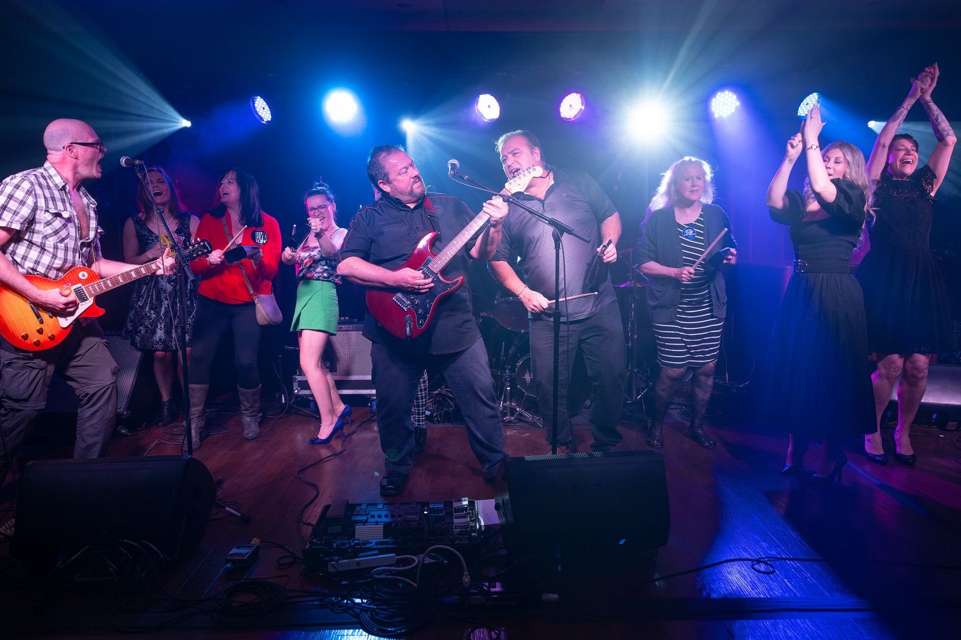 Band performing on stage, lit with blue and white lights. Guitarists, singers, and audience members are present.