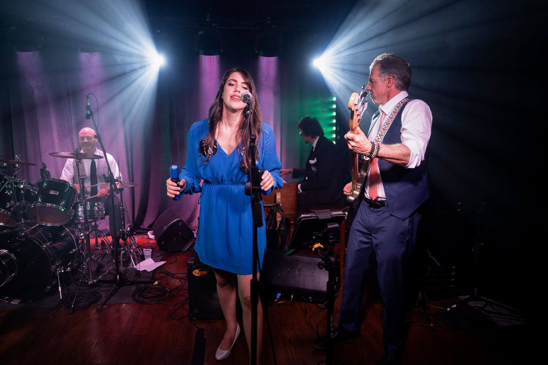 Band performing on stage: singer in blue dress, guitarist, drummer, keyboardist, spotlights.