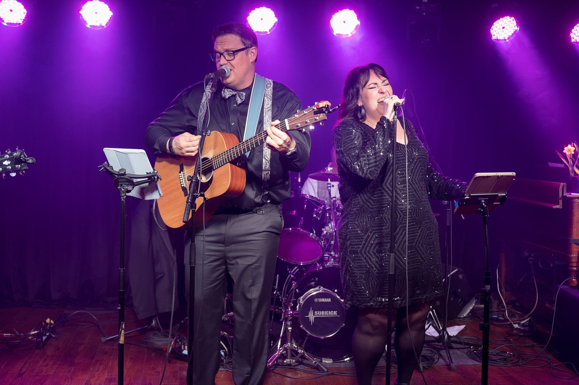 Two musicians on stage under purple lights: a guitarist with glasses, a vocalist in a sequined dress.