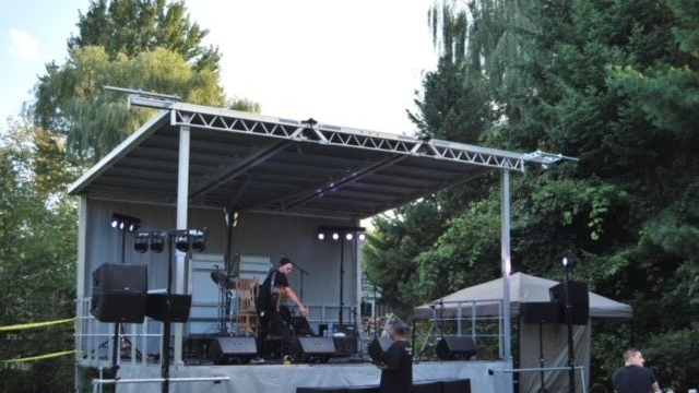 Outdoor stage set up for a performance. Person on stage, sound equipment, trees in the background.