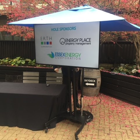 A screen displaying sponsor logos is under a blue umbrella, on a stand at an outdoor event.