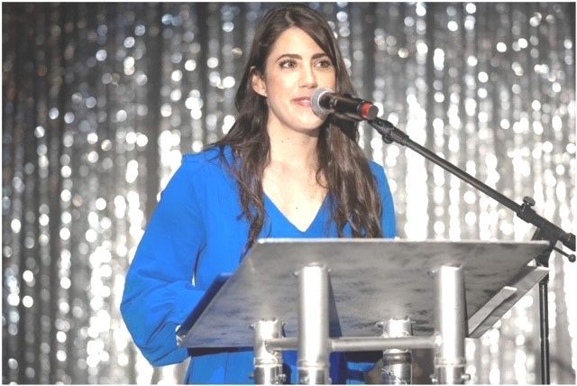 Woman speaking at a podium, wearing a blue blazer, in front of a shiny silver backdrop.