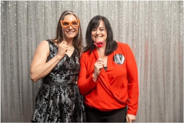 Two women pose at photo booth, one with orange glasses and the other with a red bow, smiling in front of a silver backdrop.