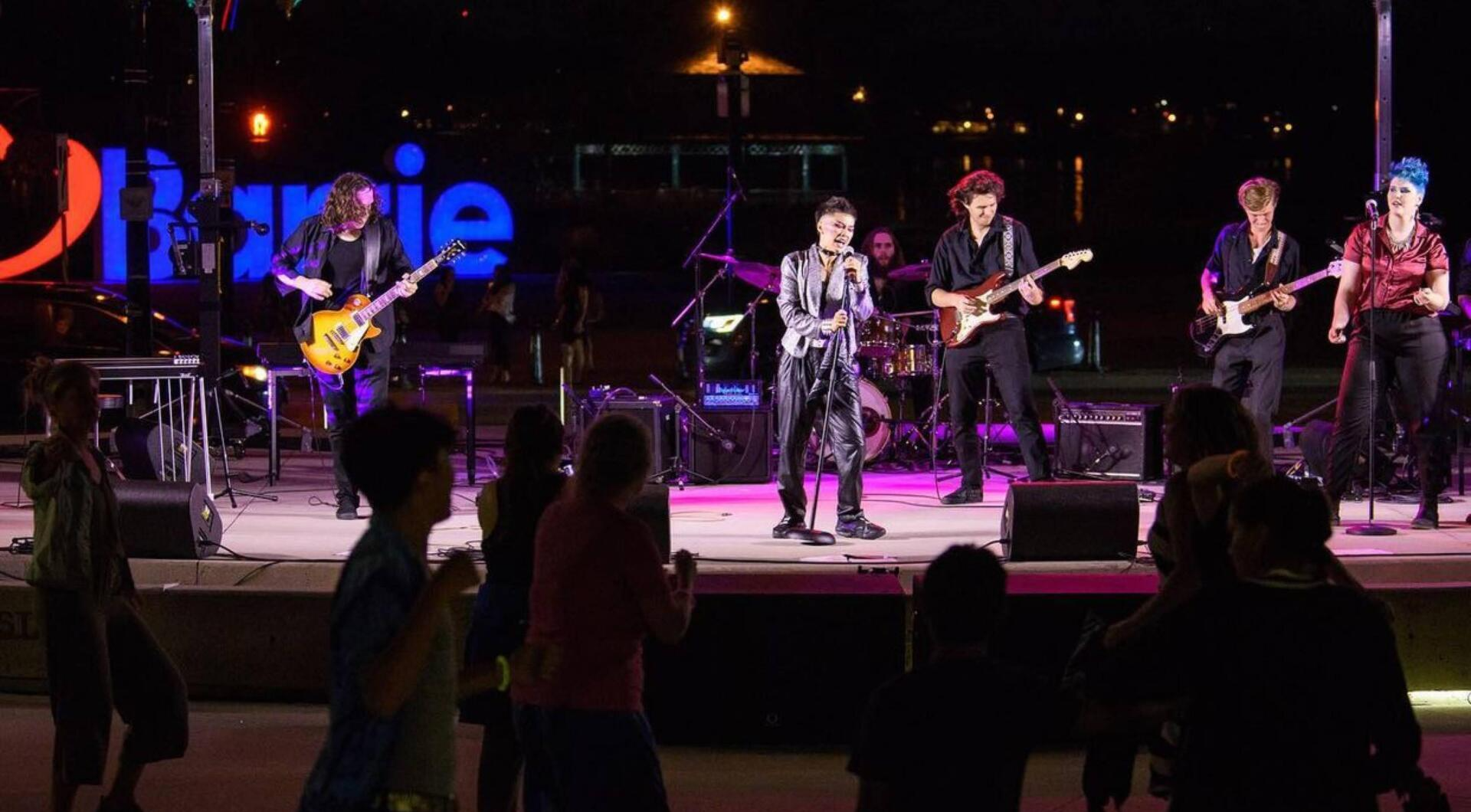 Band performing on a stage at night with audience dancing. Neon sign in background.