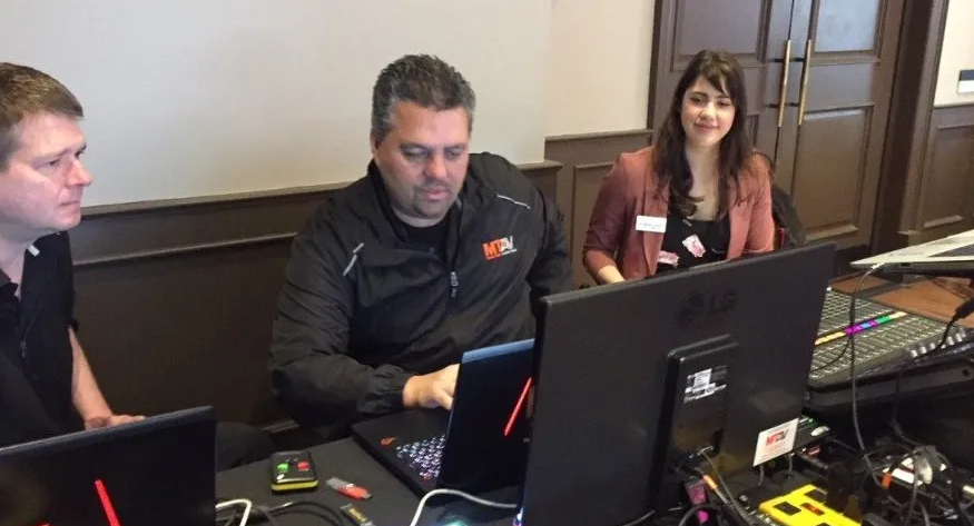 Three people at a table with laptops and a soundboard. Woman smiles while others look at screens.