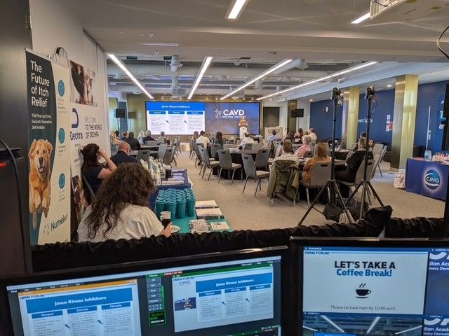 the view from the technicians table with computers set up. there are attendees sitting in the chairs and a blue and white sceen at the front.