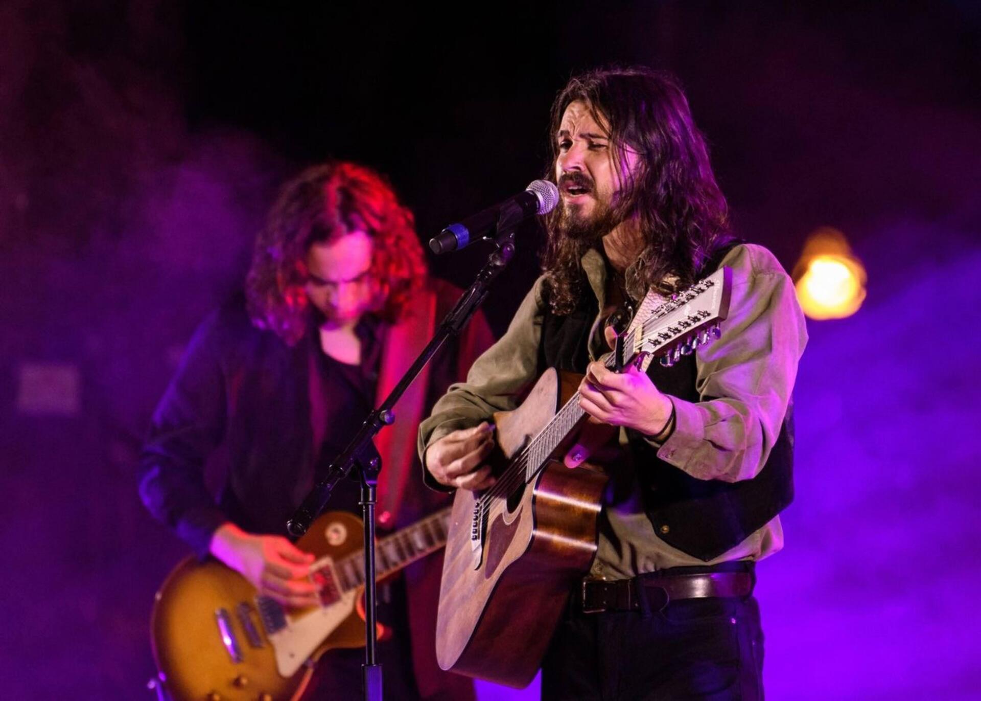 Two musicians performing on stage: one with acoustic guitar, the other with electric guitar. Purple lighting and smoke.