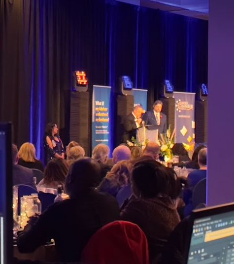 People seated at tables, listening to speakers on stage at an event, blue backdrop.