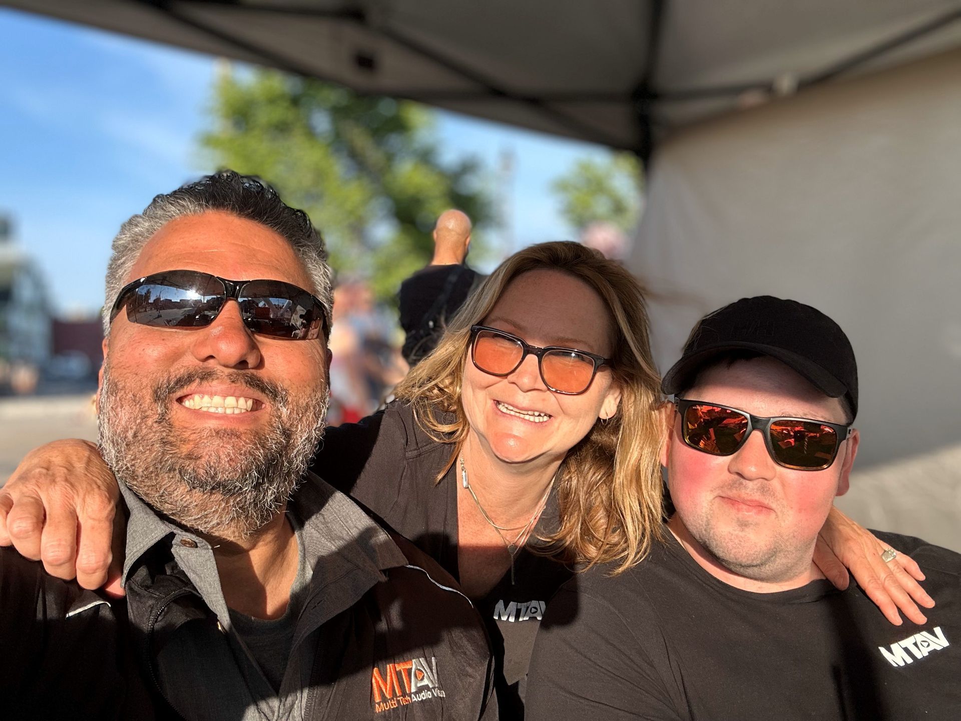 Three people smiling, wearing sunglasses, under a canopy. Outdoor daytime setting. Man on left has arm around woman.