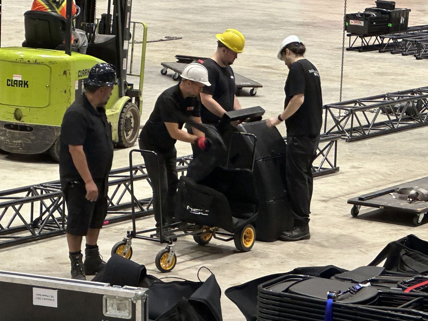 Four people with hard hats loading a black case onto a cart in a warehouse, a forklift is nearby.