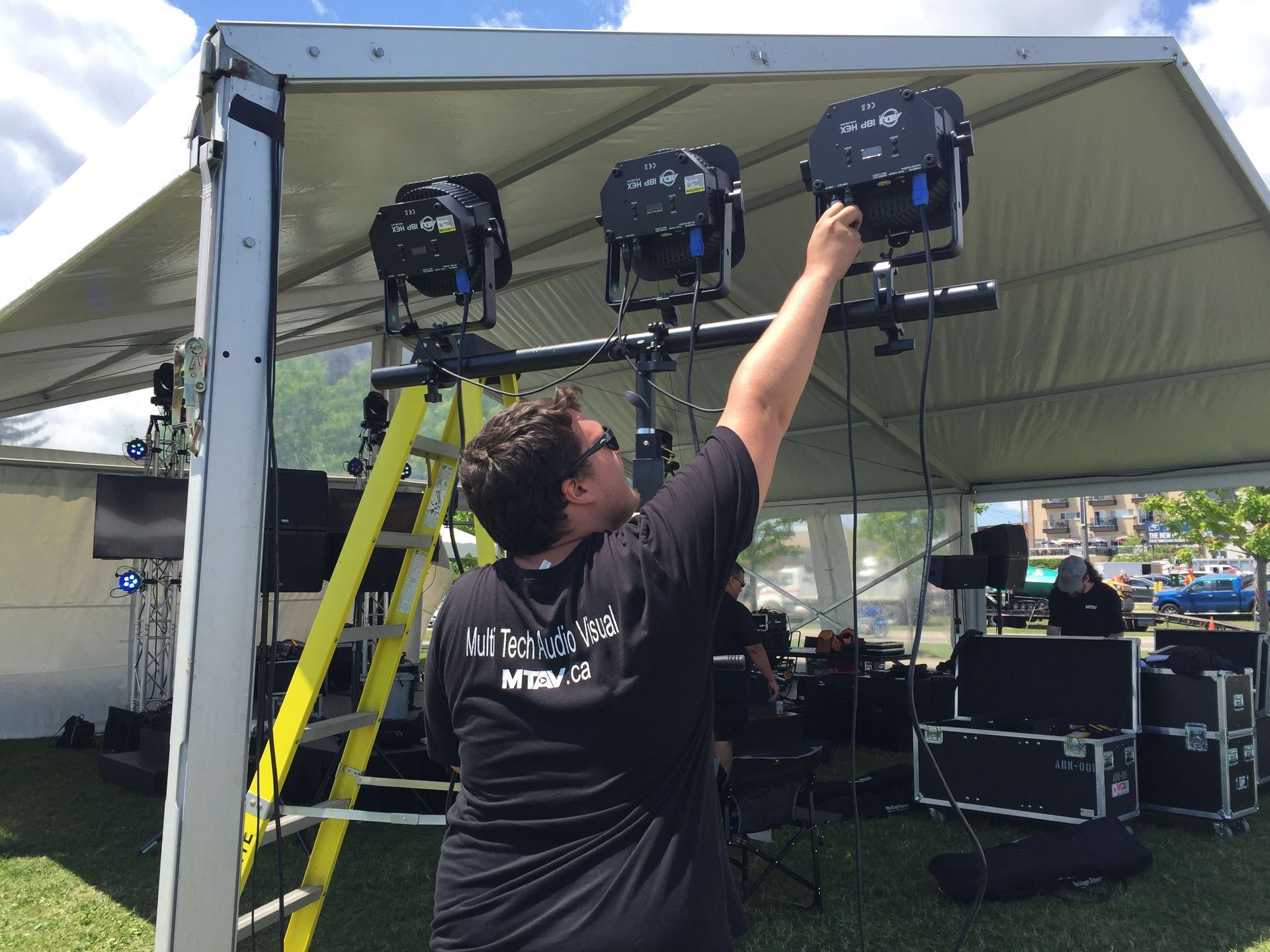 Man adjusting stage lights on a truss above a grassy outdoor event space.