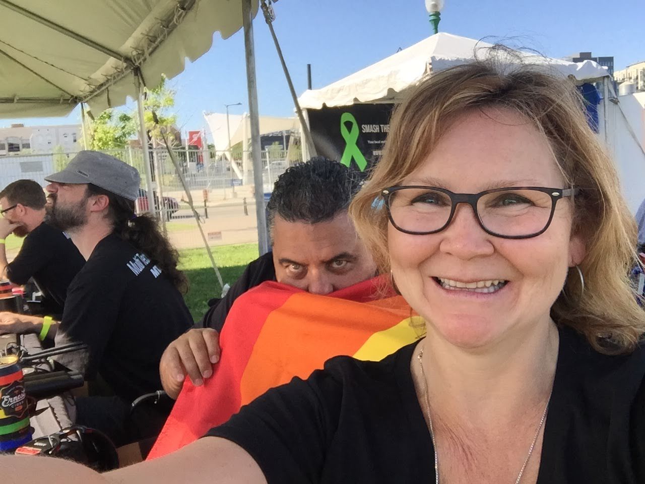 Woman smiles for selfie, man hides behind her with rainbow flag at outdoor event. Others work behind them.