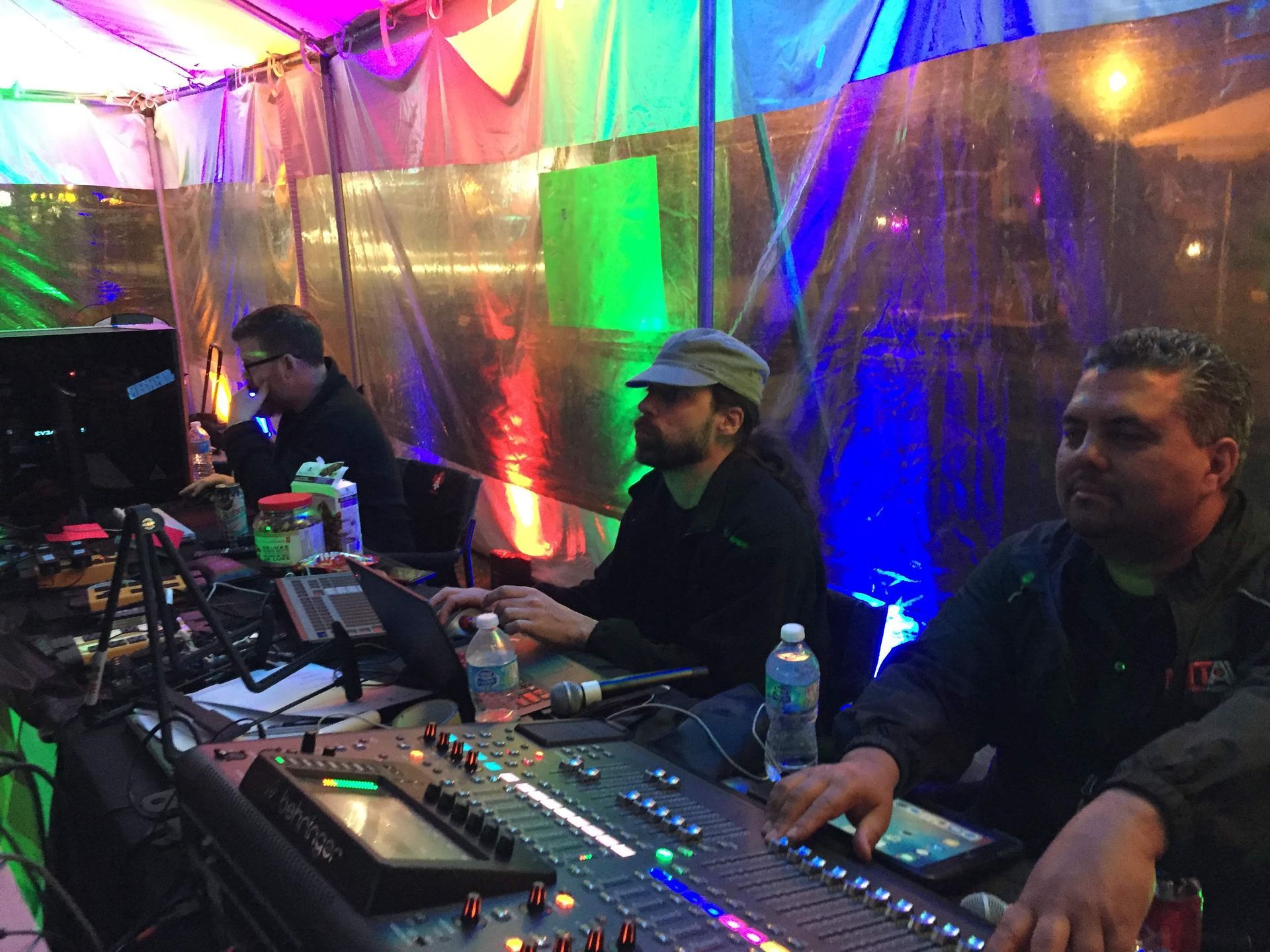 Three people operating sound equipment under a colorful canopy, outdoors at night.