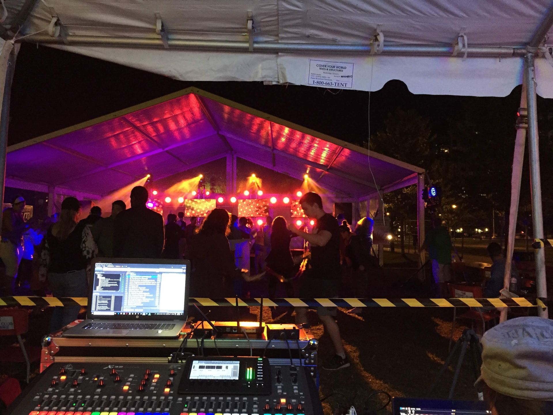 Nighttime outdoor event: People under a tent lit with purple and red lights. DJ equipment in foreground.