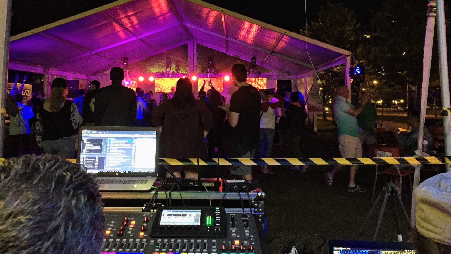 DJ setup in front of a tent with people dancing. Purple and pink lights illuminate the stage.