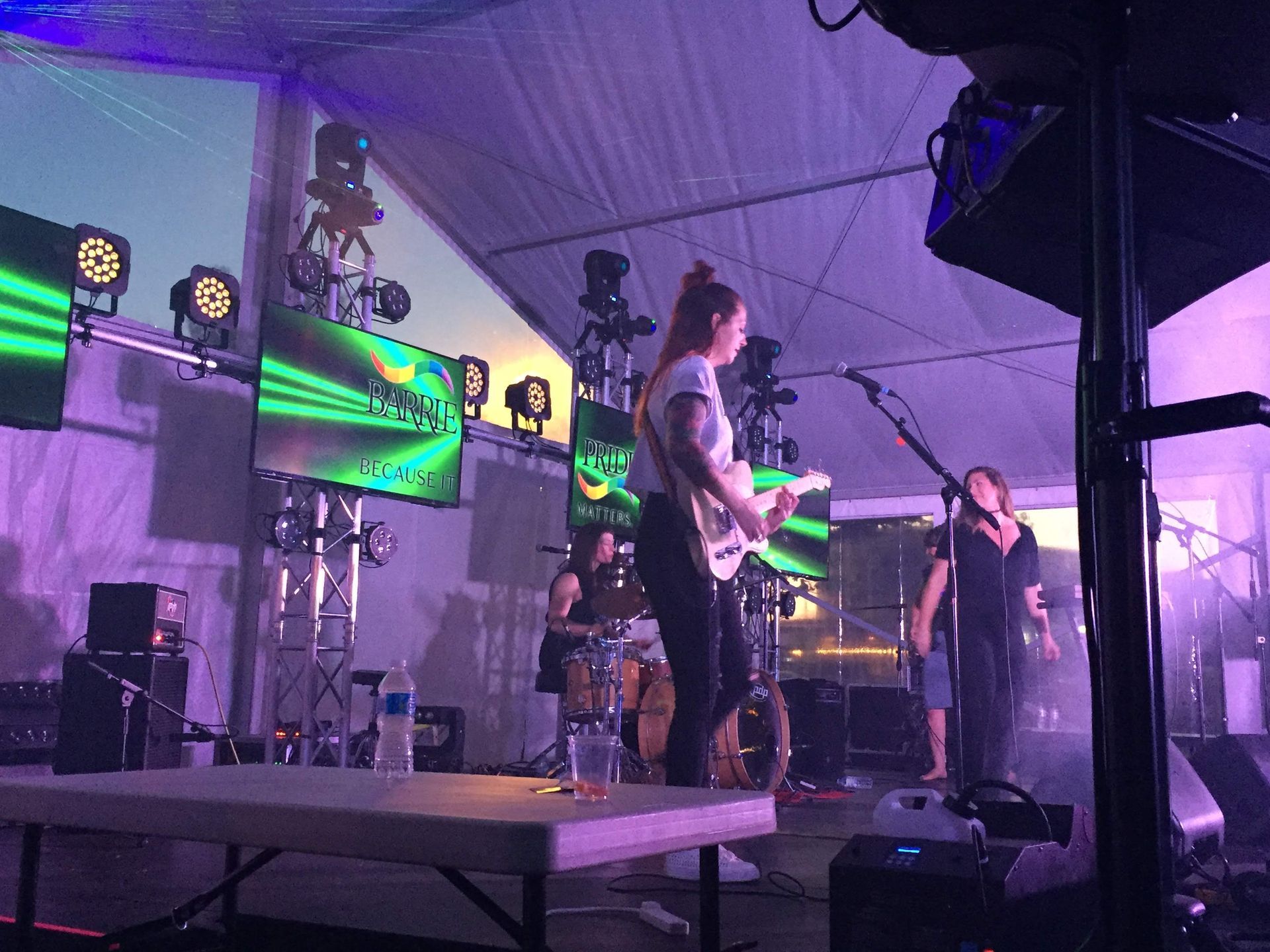 A band performs on stage under a tent, using guitars and a drum set with screen graphics. Purple stage lights.