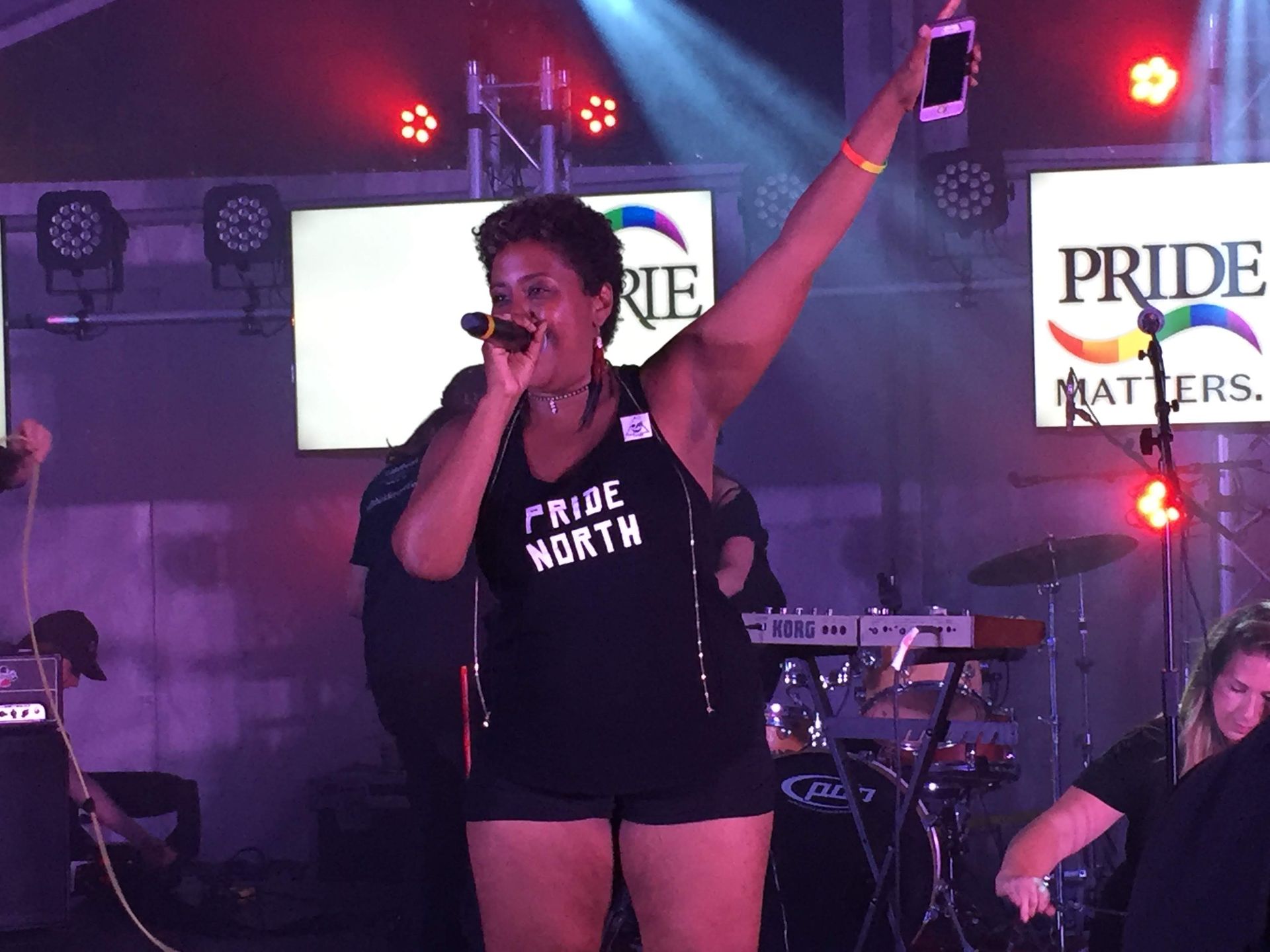 Woman singing at Pride event with raised arm holding phone. Stage with lights and 