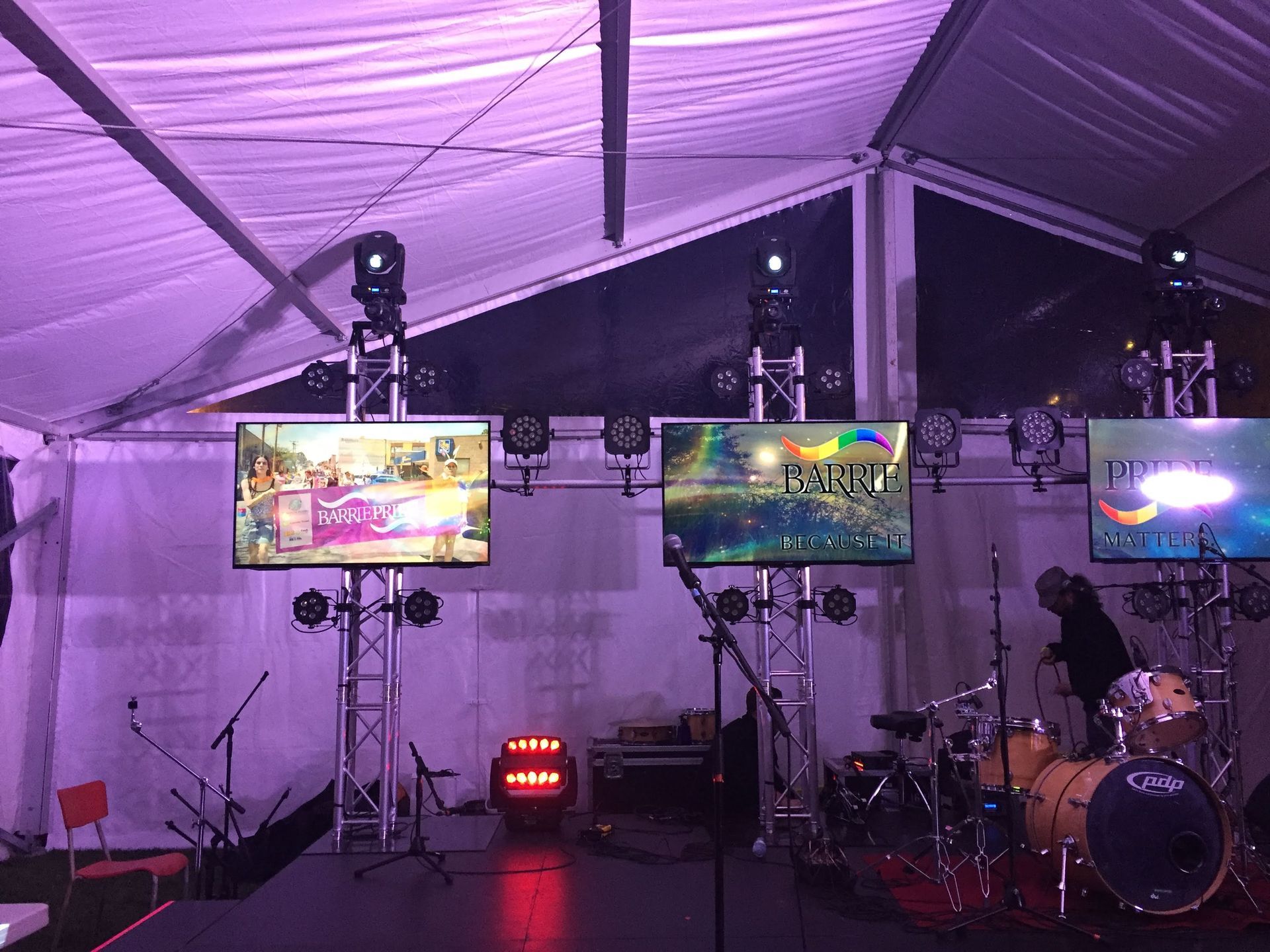 Stage setup inside a tent with three screens displaying images, lights, and a drum set.