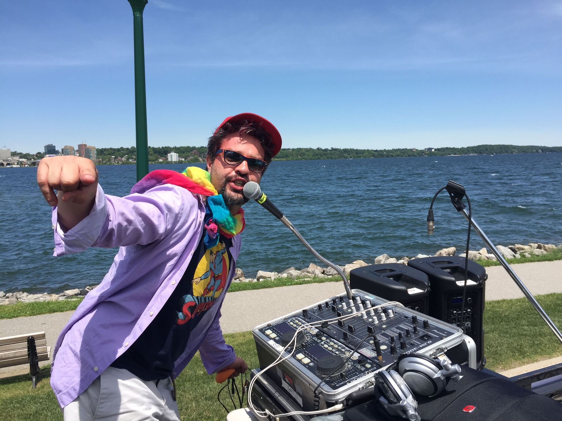 DJ with a rainbow scarf and red cap performs outdoors near a body of water, raising his fist.
