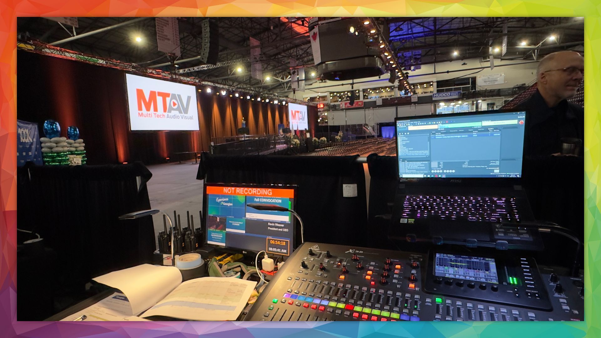 Sound engineer's workstation at a conference. Laptop, mixing board, monitors, and stage in the background.