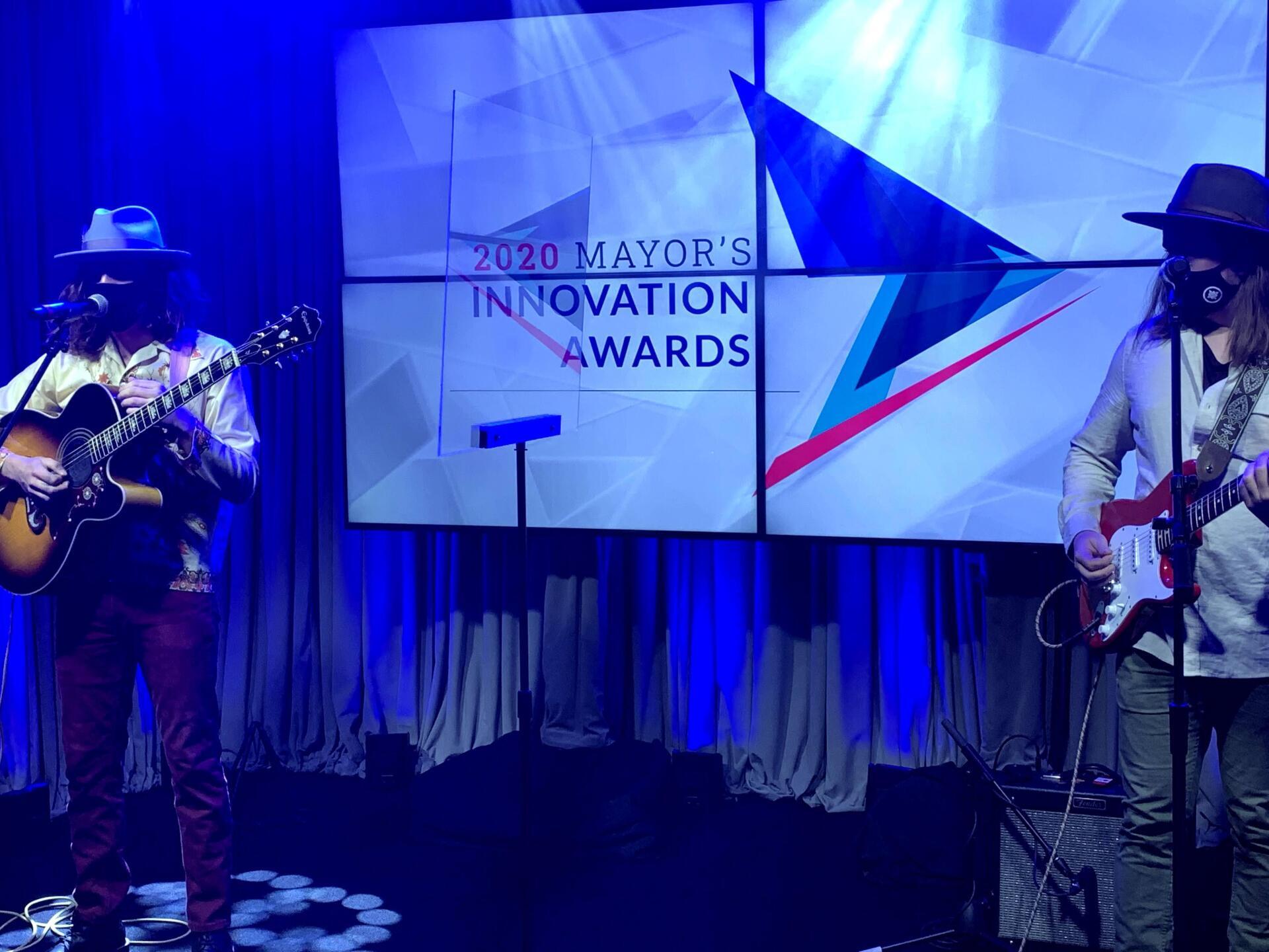 Two musicians with hats and guitars on stage with a screen behind them that says 