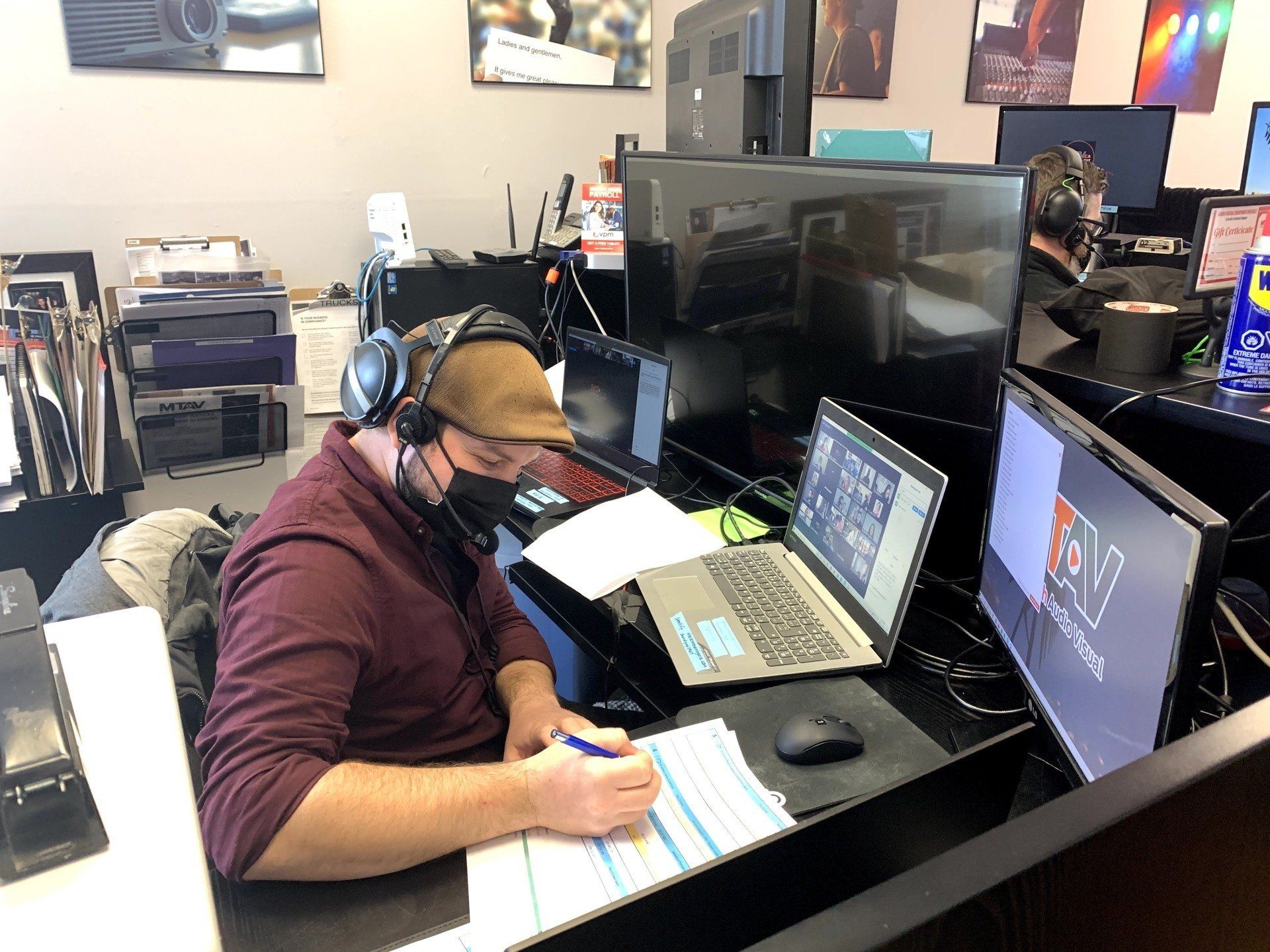 Man wearing headphones and a mask works at a desk with multiple monitors, writing on a document.