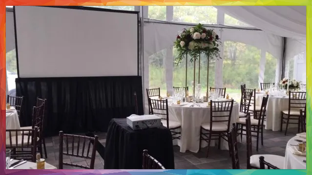 Wedding reception in a tent with round tables, floral centerpiece, and projector screen.