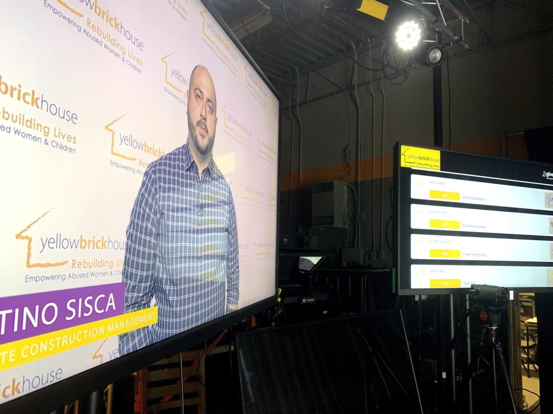 Man on a screen behind a set, promoting Yellow Brick House, with teleprompter in foreground.