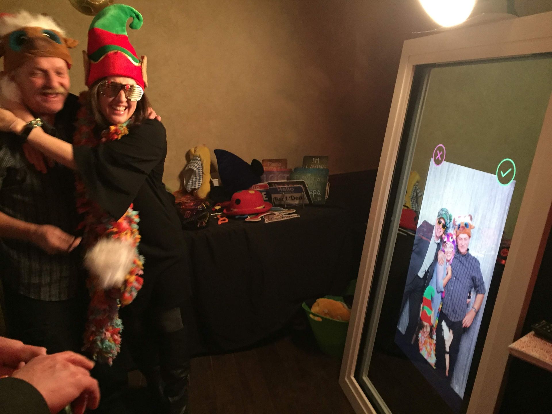 Two people wearing elf hats pose in front of a photo booth mirror, smiling and embracing.