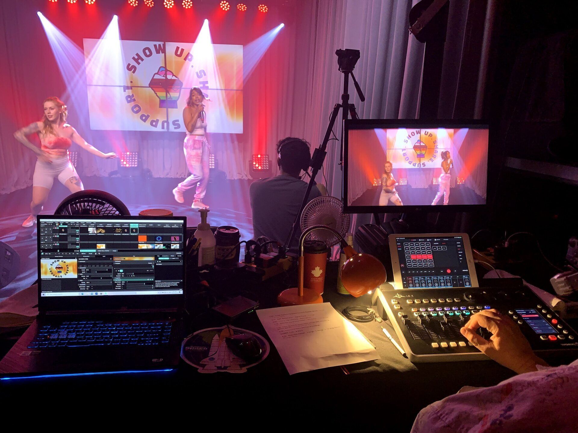 Stage production with dancers, monitors, and sound equipment. Dancers on stage, show title displayed on screens.