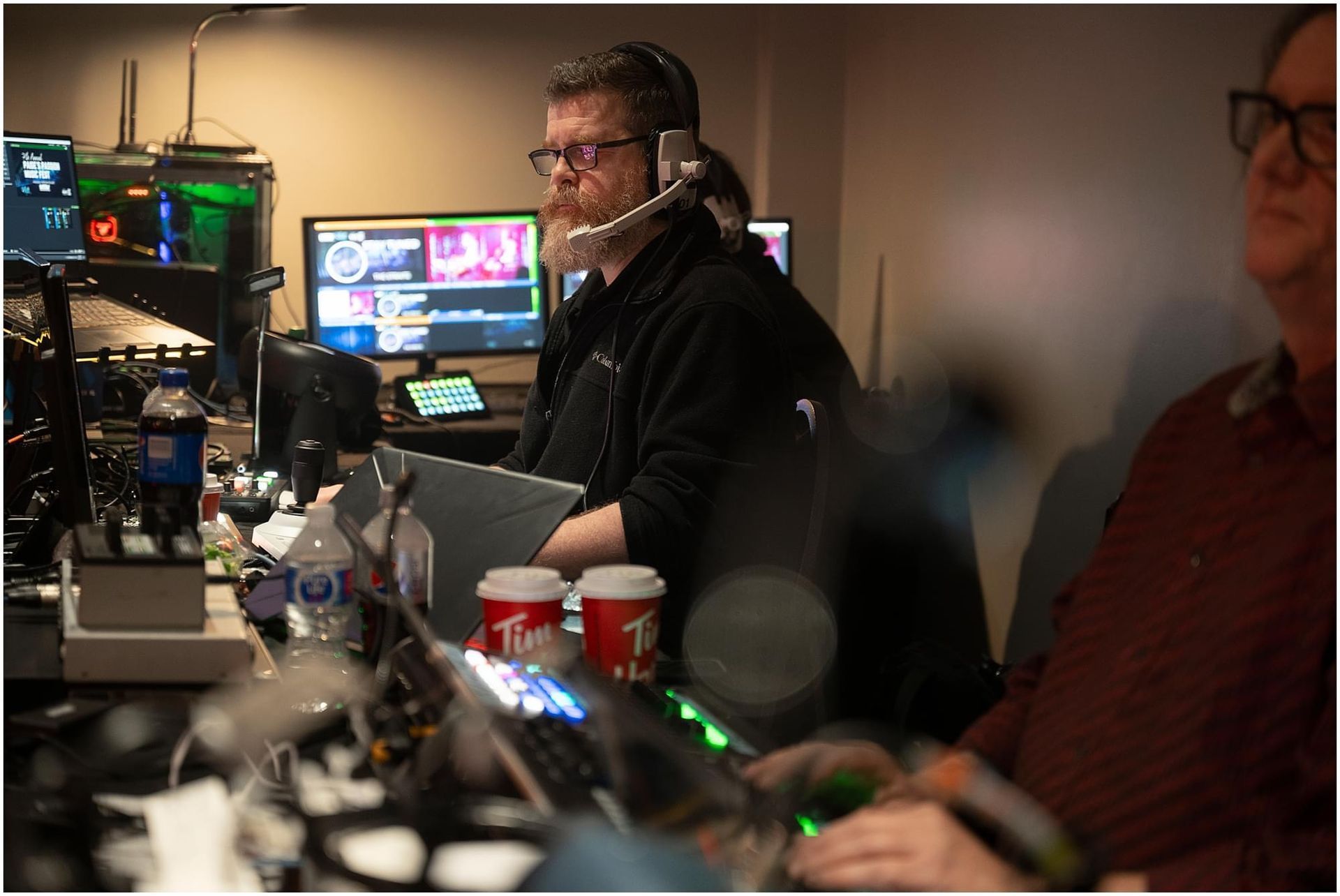 Man in headset operates control panel, monitors with live feeds. Coffee cups and computer equipment visible.