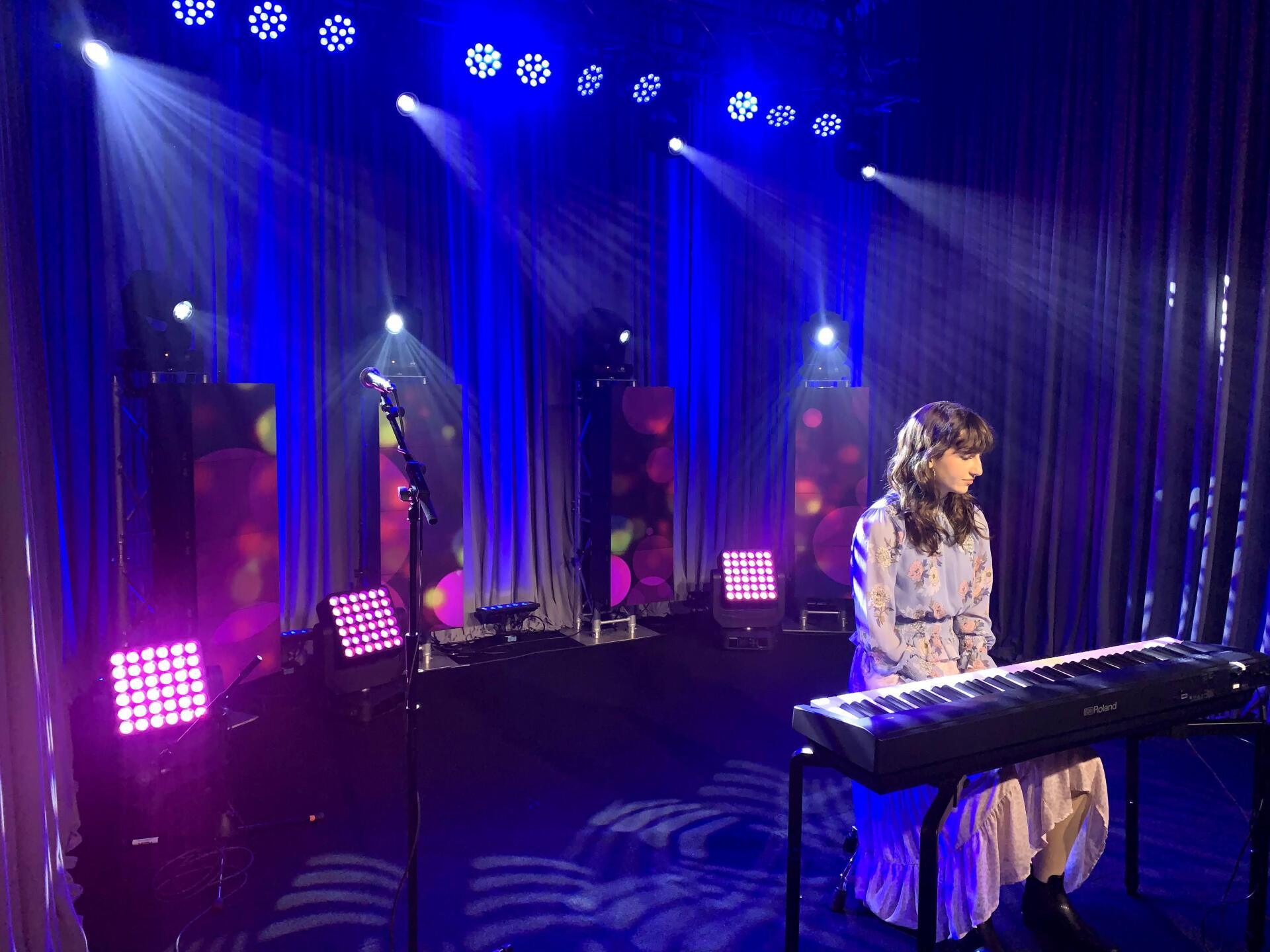 Woman playing keyboard on a stage with blue lighting and curtains.