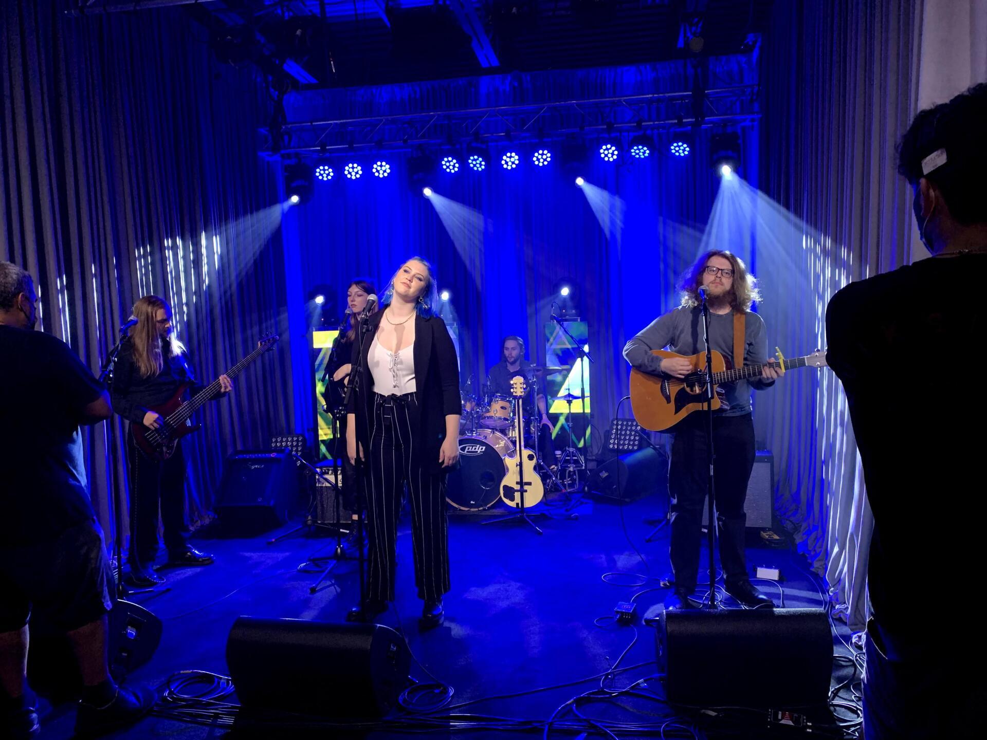 Band performing on a stage lit with blue lights. Musicians play instruments, singing into microphones.