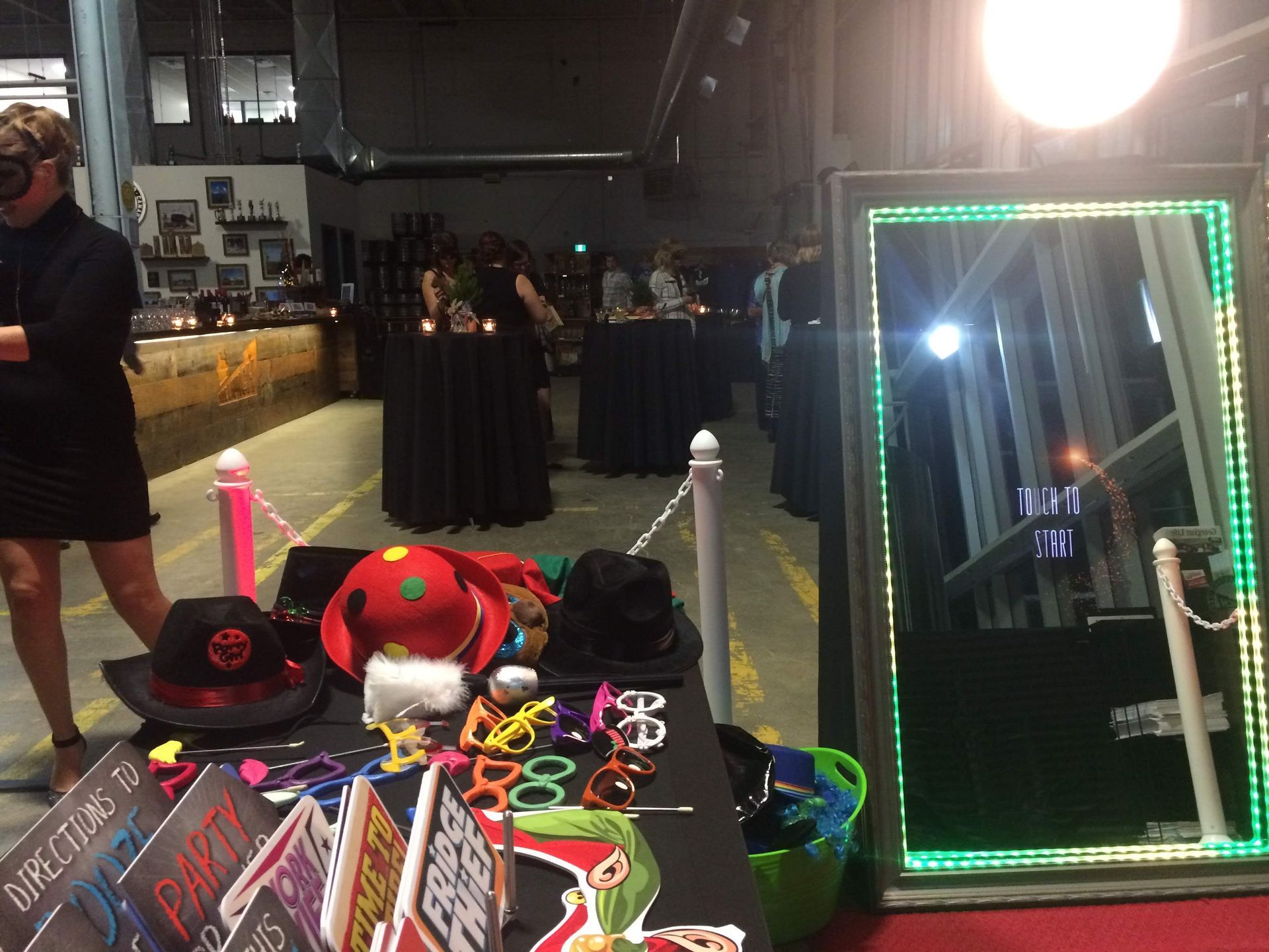 Party with a photo booth; a mirror with green lights, accessories on a table, and people at tables.
